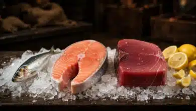 Different fish on crushed ice at a market — a small sardine, a salmon steak, and a large tuna steak, with lemons beside them.
