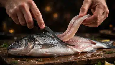 Hands filleting a whole fresh fish on a wooden board with a flexible fillet knife, one fillet lifting away.