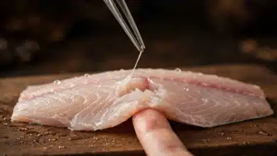 Fish tweezers pulling a thin pin bone from a raw fish fillet on a wooden board, a fingertip holding the flesh steady.